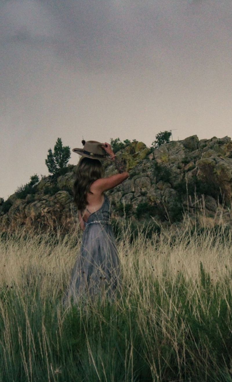 Kaylie Michelle in a grassy field, seen from behind, holding the brim of her hat